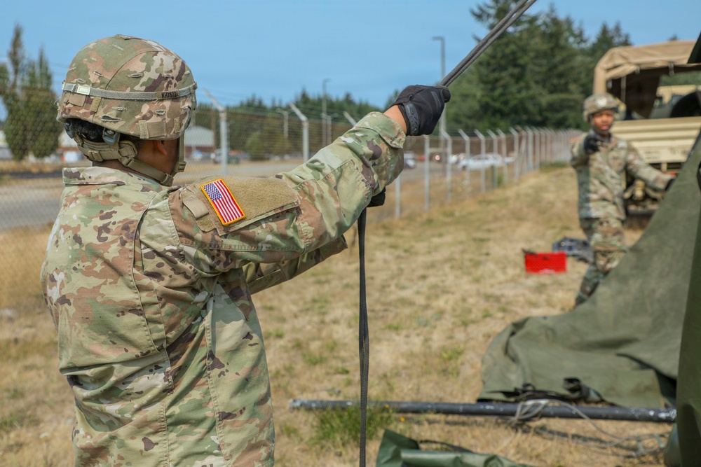 Two U.S. Army soldiers in Operational Camouflage Pattern (OCP) uniforms are conducting field equipment setup operations outdoors. The foreground soldier is handling a cable or antenna mast while wearing tactical gloves and a helmet with communications equipment. A second soldier is visible in the background near a military vehicle, and green canvas equipment is partially visible in the foreground.