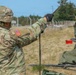 Two U.S. Army soldiers in Operational Camouflage Pattern (OCP) uniforms are conducting field equipment setup operations outdoors. The foreground soldier is handling a cable or antenna mast while wearing tactical gloves and a helmet with communications equipment. A second soldier is visible in the background near a military vehicle, and green canvas equipment is partially visible in the foreground.