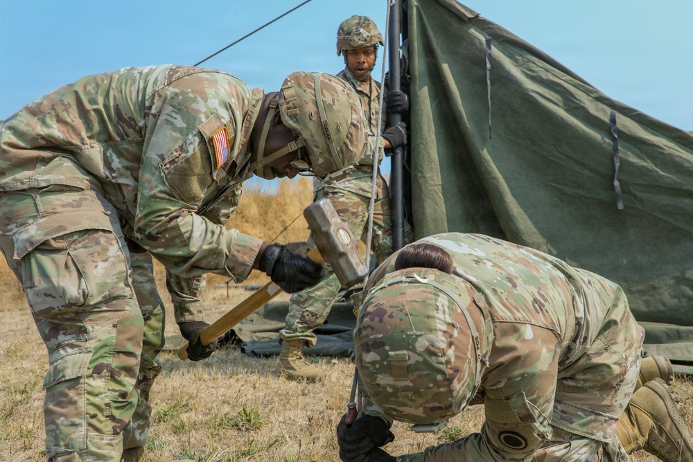 Three U.S. Army soldiers in OCP (Operational Camouflage Pattern) uniforms are setting up a military field tent on an open grassy area. One soldier is using a mallet to drive a tent stake into the ground while another crouches nearby holding the stake, and a third soldier stands holding a tent pole in the background. The scene appears to be a field training exercise or forward operating base establishment.