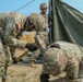 Three U.S. Army soldiers in OCP (Operational Camouflage Pattern) uniforms are setting up a military field tent on an open grassy area. One soldier is using a mallet to drive a tent stake into the ground while another crouches nearby holding the stake, and a third soldier stands holding a tent pole in the background. The scene appears to be a field training exercise or forward operating base establishment.