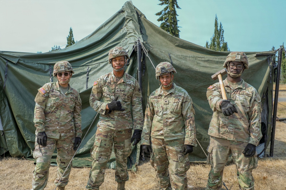 Four U.S. Army soldiers in Operational Camouflage Pattern (OCP) uniforms pose together in front of a large military field tent. They are wearing combat helmets and tactical gloves, with one soldier on the far right holding a mallet or sledgehammer. The setting appears to be a field training exercise or base camp environment surrounded by conifer trees.
