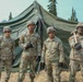 Four U.S. Army soldiers in Operational Camouflage Pattern (OCP) uniforms pose together in front of a large military field tent. They are wearing combat helmets and tactical gloves, with one soldier on the far right holding a mallet or sledgehammer. The setting appears to be a field training exercise or base camp environment surrounded by conifer trees.