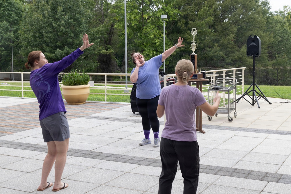 Three women are gathered on an outdoor paved plaza, appearing to celebrate or react enthusiastically with raised arms and laughter. The central figure in a light blue shirt is laughing with both arms raised, while the other two women mirror similar gestures. A trophy, speaker on a tripod, and a utility cart are visible in the background, suggesting a recent event or competition.