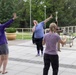 Three women are gathered on an outdoor paved plaza, appearing to celebrate or react enthusiastically with raised arms and laughter. The central figure in a light blue shirt is laughing with both arms raised, while the other two women mirror similar gestures. A trophy, speaker on a tripod, and a utility cart are visible in the background, suggesting a recent event or competition.