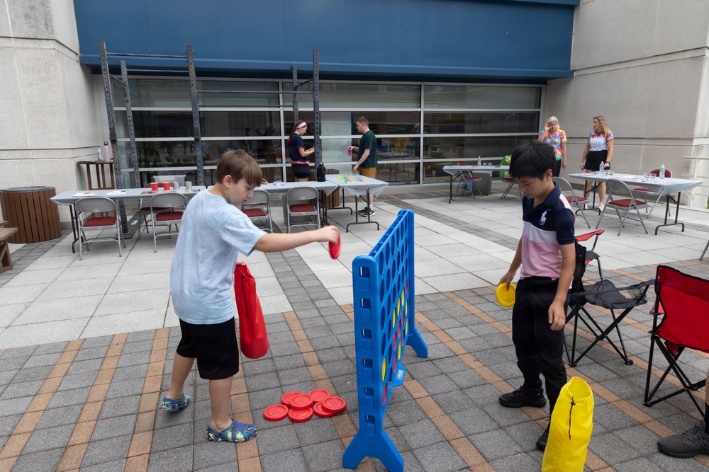 Two children are playing a giant Connect Four game outdoors on a paved plaza area. One child holds red discs while the other holds yellow discs, actively engaged in gameplay. In the background, several adults are gathered around tables, suggesting an organized outdoor event or activity fair.