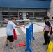 Two children are playing a giant Connect Four game outdoors on a paved plaza area. One child holds red discs while the other holds yellow discs, actively engaged in gameplay. In the background, several adults are gathered around tables, suggesting an organized outdoor event or activity fair.