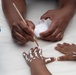 An adult is using a fine-tipped paintbrush to apply white face/body paint onto a child's hand, creating a skeletal bone pattern. The adult holds a small container of white paint in one hand while carefully painting the child's fingers and hand dorsum. The scene appears to be a craft or face-painting activity at an event or gathering.