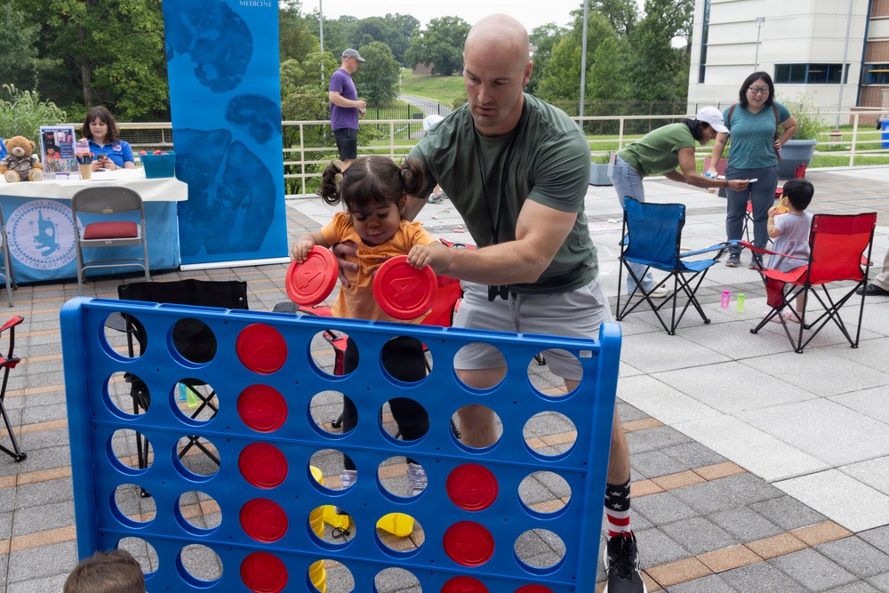 An outdoor community event where a bald adult male in a green t-shirt is helping a young toddler girl in an orange outfit play a giant Connect Four game. The event appears to be held on a paved plaza area with various activity stations, folding chairs, and informational tables visible in the background. Multiple families with children are participating in the event.