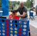 An outdoor community event where a bald adult male in a green t-shirt is helping a young toddler girl in an orange outfit play a giant Connect Four game. The event appears to be held on a paved plaza area with various activity stations, folding chairs, and informational tables visible in the background. Multiple families with children are participating in the event.