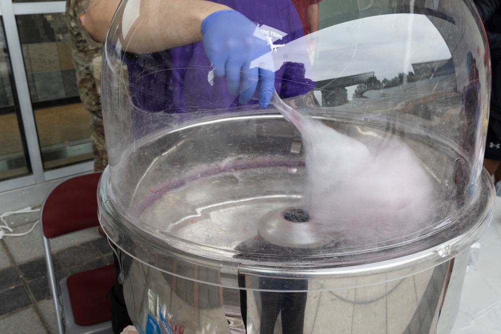 A person wearing blue latex gloves is making cotton candy using a commercial cotton candy machine with a clear dome cover. Pink/purple spun sugar strands are visible forming inside the stainless steel spinning bowl. The setting appears to be an outdoor or semi-outdoor event.