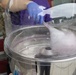A person wearing blue latex gloves is making cotton candy using a commercial cotton candy machine with a clear dome cover. Pink/purple spun sugar strands are visible forming inside the stainless steel spinning bowl. The setting appears to be an outdoor or semi-outdoor event.