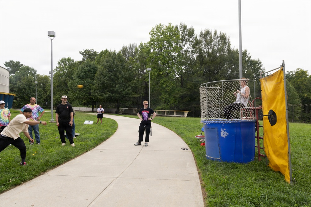 An outdoor community or school event featuring a dunk tank on the right side, where a person is seated on the platform above a large blue water barrel. On the left side, participants are playing cornhole (bean bag toss), with one person mid-throw. The setting is a paved pathway surrounded by green grass and trees under an overcast sky.
