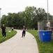 An outdoor community or school event featuring a dunk tank on the right side, where a person is seated on the platform above a large blue water barrel. On the left side, participants are playing cornhole (bean bag toss), with one person mid-throw. The setting is a paved pathway surrounded by green grass and trees under an overcast sky.