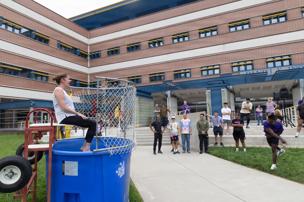 An outdoor event is taking place in front of a multi-story brick building partially labeled 'Daniel K. In...' (likely Daniel K. Inouye). A woman in a white tank top and black pants is seated on a dunk tank platform above a large blue water tank, while a person on the right side appears to be throwing a ball to trigger the mechanism. A crowd of approximately 15-20 spectators watches from the surrounding area.