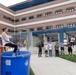 An outdoor event is taking place in front of a multi-story brick building partially labeled 'Daniel K. In...' (likely Daniel K. Inouye). A woman in a white tank top and black pants is seated on a dunk tank platform above a large blue water tank, while a person on the right side appears to be throwing a ball to trigger the mechanism. A crowd of approximately 15-20 spectators watches from the surrounding area.