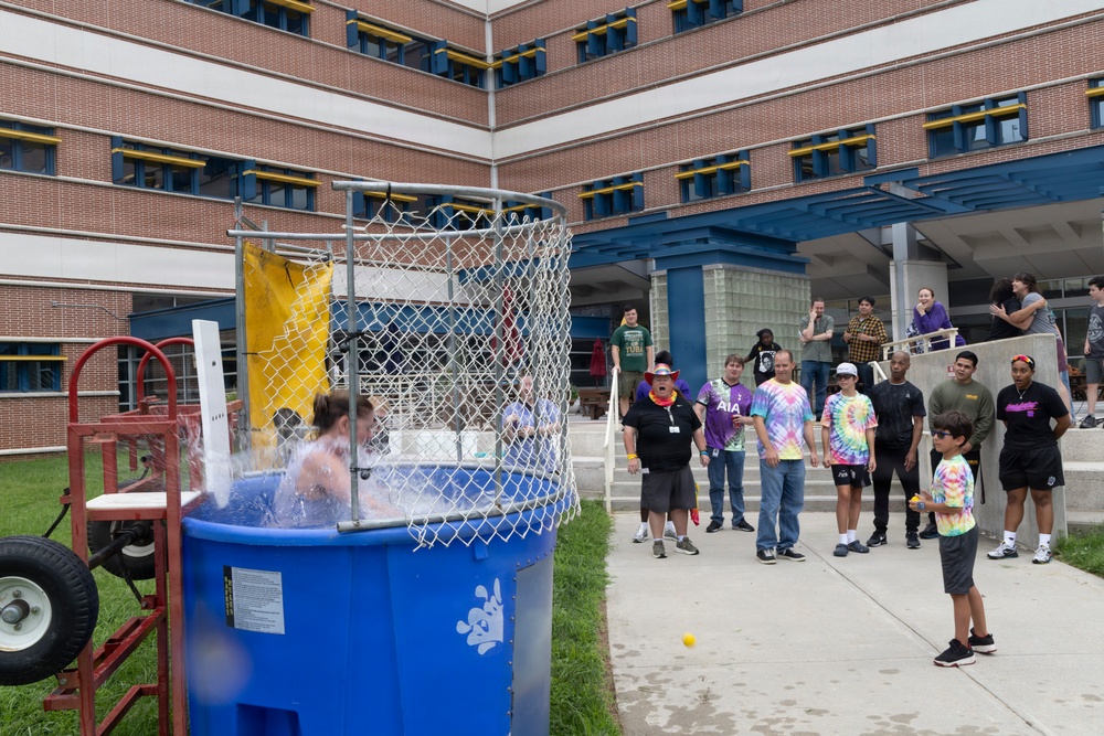 An outdoor dunk tank event is taking place in what appears to be a school or institutional courtyard. A person has just been dunked into the blue water tank, visible splashing water, while a crowd of spectators watches from the sidewalk. The event appears to be a school or community fundraiser or carnival activity.