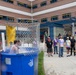 An outdoor dunk tank event is taking place in what appears to be a school or institutional courtyard. A person has just been dunked into the blue water tank, visible splashing water, while a crowd of spectators watches from the sidewalk. The event appears to be a school or community fundraiser or carnival activity.