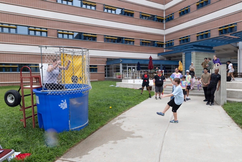 An outdoor community or workplace event is taking place in the courtyard of a multi-story brick building. A child is throwing a ball at a dunk tank, where a person is seated inside the cage over a large blue water tank. A group of spectators, including adults and children, are watching the activity from the surrounding area.