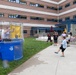 An outdoor community or workplace event is taking place in the courtyard of a multi-story brick building. A child is throwing a ball at a dunk tank, where a person is seated inside the cage over a large blue water tank. A group of spectators, including adults and children, are watching the activity from the surrounding area.