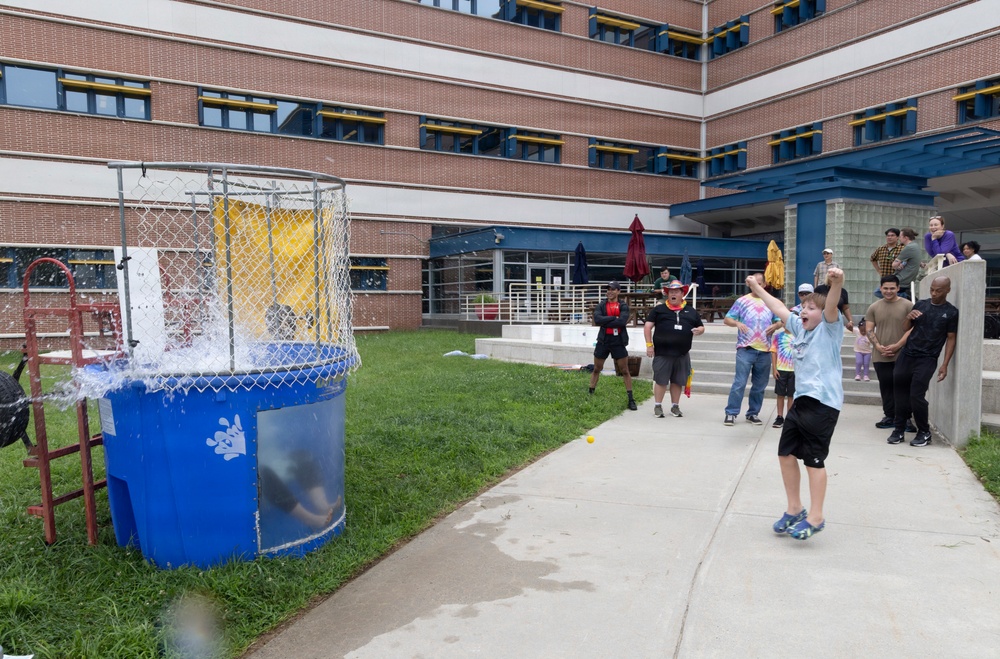 An outdoor event featuring a dunk tank is taking place in the courtyard of a multi-story brick building. A child wearing a tie-dye shirt is celebrating with raised arms, apparently having just successfully triggered the dunk tank, as evidenced by the water splashing inside the tank enclosure. A group of onlookers and participants are gathered nearby watching the activity.