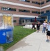 An outdoor event featuring a dunk tank is taking place in the courtyard of a multi-story brick building. A child wearing a tie-dye shirt is celebrating with raised arms, apparently having just successfully triggered the dunk tank, as evidenced by the water splashing inside the tank enclosure. A group of onlookers and participants are gathered nearby watching the activity.