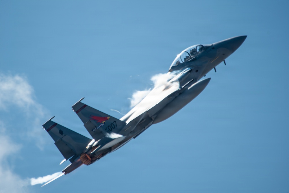 A military jet fighter is captured in a steep climbing maneuver against a blue sky, with afterburners visibly ignited and condensation vapor trails forming over the wings. The aircraft bears the tail number '007' and displays a red insignia patch on the fuselage, consistent with a Republic of Korea Air Force (ROKAF) F-15K Eagle. The image is sharp and well-composed, clearly showing the aircraft's details during a high-performance flight demonstration.