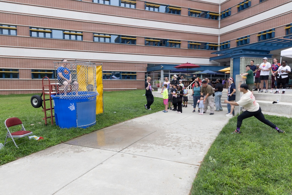 An outdoor community or workplace event featuring a dunk tank activity in front of a multi-story brick building. A person in a blue shirt is seated on the dunk tank platform while another person in a white shirt is in a throwing stance, preparing to hit the target. A crowd of spectators of various ages watches from the sidewalk and steps nearby.