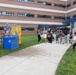 An outdoor community or workplace event featuring a dunk tank activity in front of a multi-story brick building. A person in a blue shirt is seated on the dunk tank platform while another person in a white shirt is in a throwing stance, preparing to hit the target. A crowd of spectators of various ages watches from the sidewalk and steps nearby.