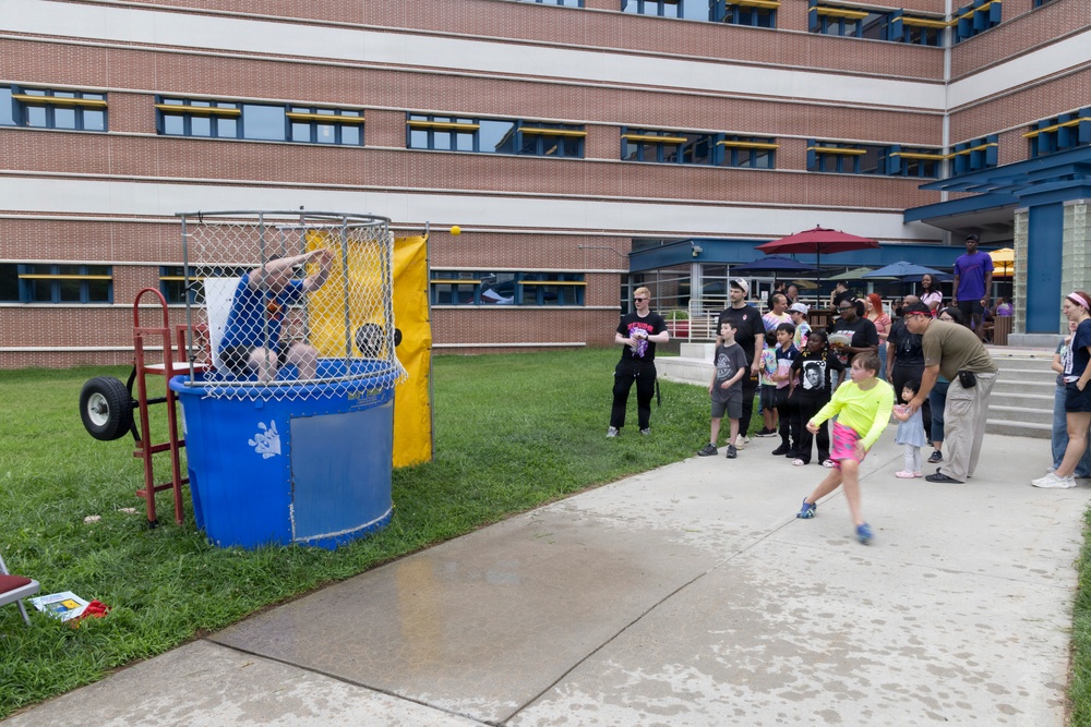 An outdoor community or school event featuring a dunk tank activity. A child in a neon yellow-green shirt is in the act of throwing a ball at the dunk tank target, while a person is already submerged or falling inside the blue tank. A crowd of adults and children watches from the sideline near a brick building.