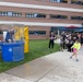 An outdoor community or school event featuring a dunk tank activity. A child in a neon yellow-green shirt is in the act of throwing a ball at the dunk tank target, while a person is already submerged or falling inside the blue tank. A crowd of adults and children watches from the sideline near a brick building.