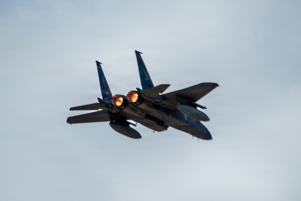A twin-engine military fighter jet, consistent with an F-15 Eagle, is photographed in flight against an overcast sky. The aircraft is viewed from a rear-quarter angle, with both afterburners visibly ignited, producing bright orange flames. The jet appears to be in a slight bank or climbing maneuver.