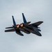 A twin-engine military fighter jet, consistent with an F-15 Eagle, is photographed in flight against an overcast sky. The aircraft is viewed from a rear-quarter angle, with both afterburners visibly ignited, producing bright orange flames. The jet appears to be in a slight bank or climbing maneuver.
