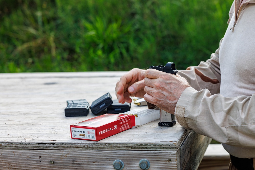 A person is at an outdoor shooting range, loading ammunition into a firearm magazine on a wooden bench. A red box of Federal .40 caliber brass ammunition (50 rounds, 180 grain) is open on the table alongside multiple loaded and unloaded magazines. The setting appears to be a casual range session in a vegetated outdoor environment.