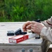 A person is at an outdoor shooting range, loading ammunition into a firearm magazine on a wooden bench. A red box of Federal .40 caliber brass ammunition (50 rounds, 180 grain) is open on the table alongside multiple loaded and unloaded magazines. The setting appears to be a casual range session in a vegetated outdoor environment.