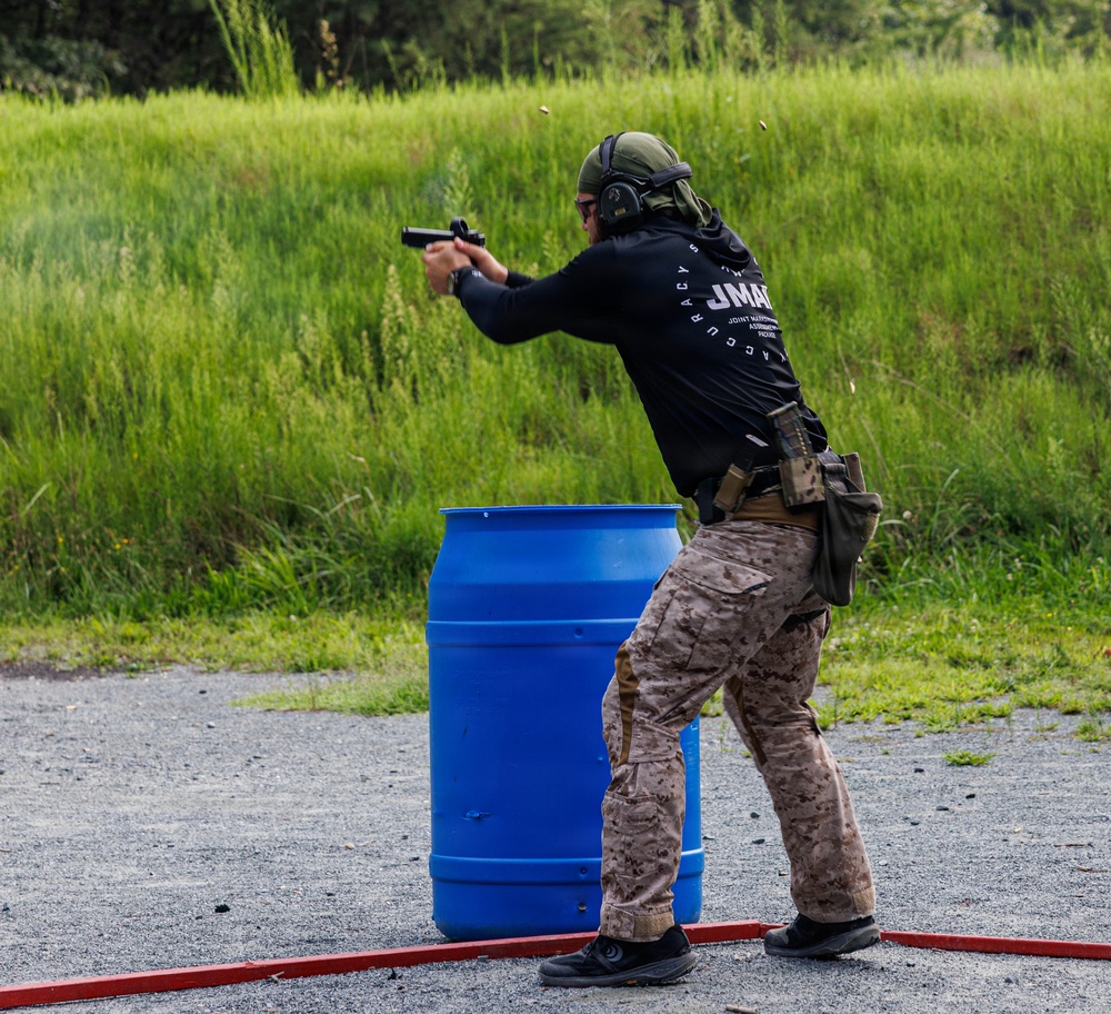 A shooter in tactical gear is firing a pistol at a shooting range, positioned behind a blue barrel used as cover. The individual is wearing a black shirt with 'JMAP' (Joint Marksmanship Assessment Program) text, desert camouflage pants, and a green helmet with ear protection. A spent casing is visibly ejecting from the firearm, indicating an active firing moment.