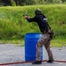 A shooter in tactical gear is firing a pistol at a shooting range, positioned behind a blue barrel used as cover. The individual is wearing a black shirt with 'JMAP' (Joint Marksmanship Assessment Program) text, desert camouflage pants, and a green helmet with ear protection. A spent casing is visibly ejecting from the firearm, indicating an active firing moment.