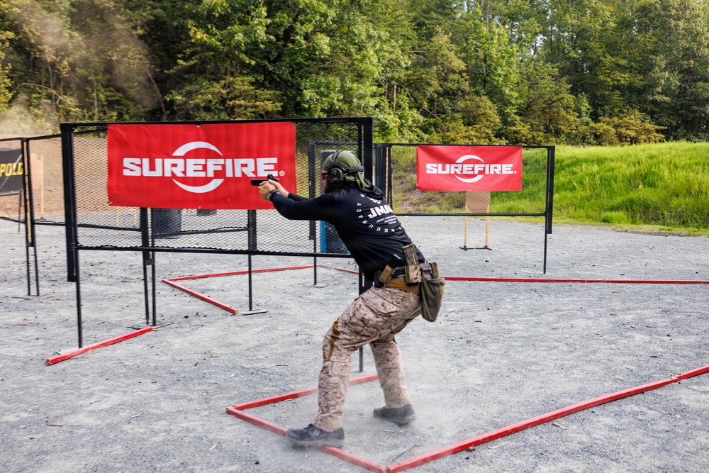 A competitive shooter is actively engaging targets at an outdoor shooting range sponsored by SureFire. The shooter is positioned within a red-bordered shooting box on a gravel surface, aiming a pistol downrange toward paper targets. The setting appears to be a structured practical shooting competition with defined stage boundaries and sponsor banners.