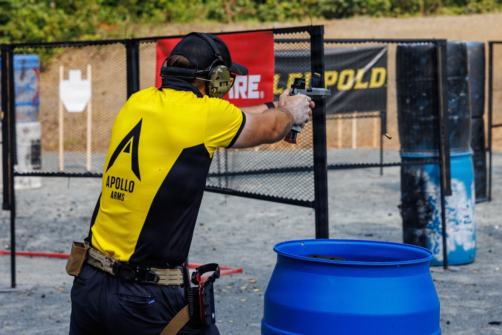 A competitive shooter wearing an 'Apollo Arms' branded yellow and black jersey is actively firing a pistol at an outdoor shooting range. A spent brass casing is visibly ejecting from the firearm, indicating the weapon has just been discharged. The setting appears to be an organized shooting competition with sponsor banners, range props, and safety equipment visible.