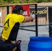 A competitive shooter wearing an 'Apollo Arms' branded yellow and black jersey is actively firing a pistol at an outdoor shooting range. A spent brass casing is visibly ejecting from the firearm, indicating the weapon has just been discharged. The setting appears to be an organized shooting competition with sponsor banners, range props, and safety equipment visible.