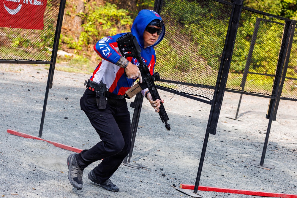 A competitive shooter is navigating a stage at what appears to be a practical shooting competition, such as IPSC or USPSA. The competitor is moving dynamically through a gate or port structure while carrying a rifle, with a holstered handgun also visible on their belt. The setting is an outdoor shooting range with gravel ground and metal stage props.
