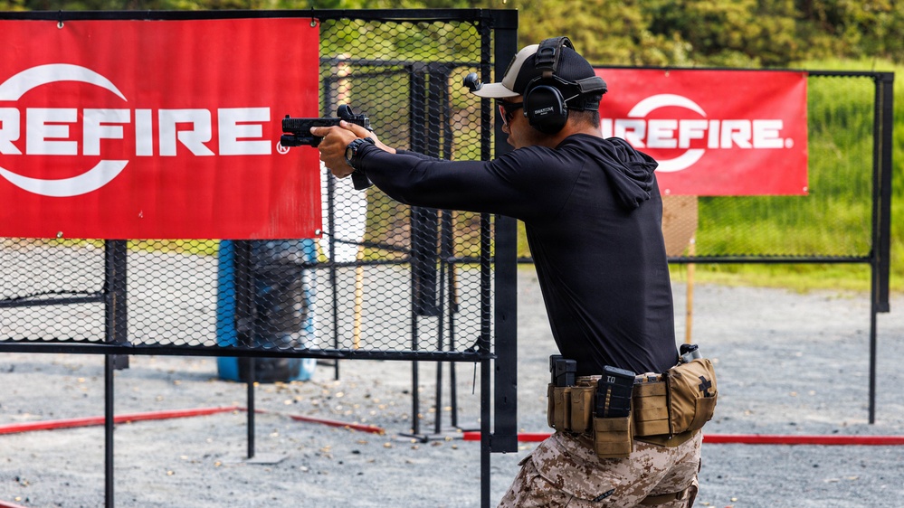A competitive shooter is seen from behind at an outdoor shooting range, aiming a pistol in a two-handed grip stance. The shooter is equipped with tactical gear including ear protection, a cap, magazine pouches on a belt, and is wearing desert camouflage pants. Red 'SureFire' branded banners are visible in the background, indicating a sponsored shooting competition or training event.