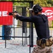 A competitive shooter is seen from behind at an outdoor shooting range, aiming a pistol in a two-handed grip stance. The shooter is equipped with tactical gear including ear protection, a cap, magazine pouches on a belt, and is wearing desert camouflage pants. Red 'SureFire' branded banners are visible in the background, indicating a sponsored shooting competition or training event.