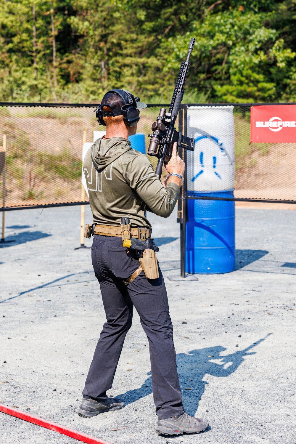 A shooter is participating in a practical shooting competition or training exercise at an outdoor range. The individual is holding a scoped AR-platform rifle in a raised, muzzle-up safe carry position while navigating the range. A 'SureFire' branded banner is visible in the background alongside target props.