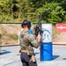 A shooter is participating in a practical shooting competition or training exercise at an outdoor range. The individual is holding a scoped AR-platform rifle in a raised, muzzle-up safe carry position while navigating the range. A 'SureFire' branded banner is visible in the background alongside target props.