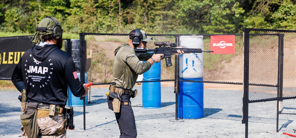 Two individuals are at an outdoor shooting range, with one actively firing a rifle equipped with an optic scope while the other observes. The setting appears to be a structured training or competition event, evidenced by sponsor signage (SureFire, Leupold) and range equipment. Both individuals are wearing tactical gear including ear protection, holsters, and caps.