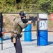 Two individuals are at an outdoor shooting range, with one actively firing a rifle equipped with an optic scope while the other observes. The setting appears to be a structured training or competition event, evidenced by sponsor signage (SureFire, Leupold) and range equipment. Both individuals are wearing tactical gear including ear protection, holsters, and caps.
