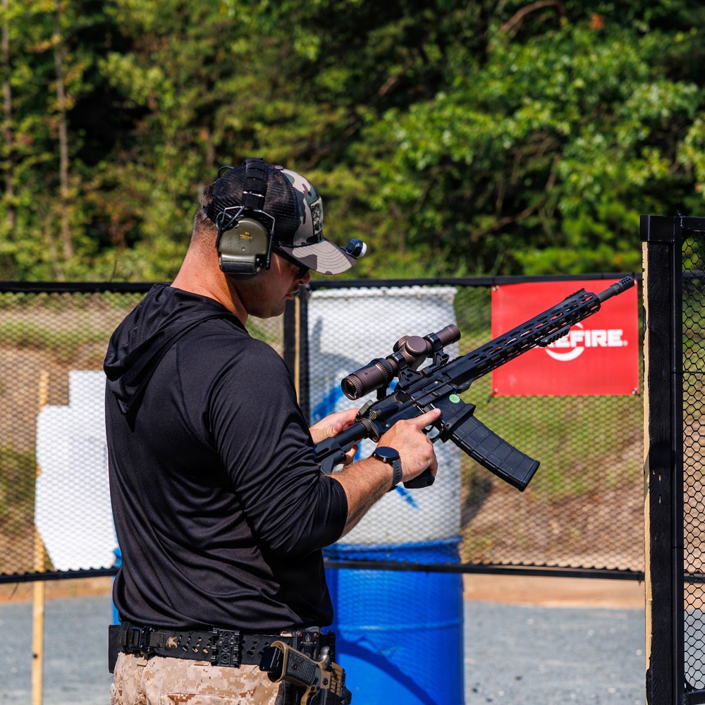 A male shooter is handling an AR-style rifle equipped with a magnified optic scope at what appears to be an organized shooting range or competition. He is wearing proper safety equipment including electronic hearing protection and a multicam baseball cap. The setting includes range props such as barrels, target stands, and a sponsor banner partially visible on the right.