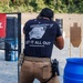 A person is actively shooting at a firearms training range, facing downrange toward silhouette targets. They are wearing tactical gear including a competition belt with holster and magazine pouches, ear protection, and a navy t-shirt reading 'LET IT ALL OUT' with the website 'financeandmaneuver.com'. A spent casing is visible in the air to the upper right, indicating the firearm was just discharged.