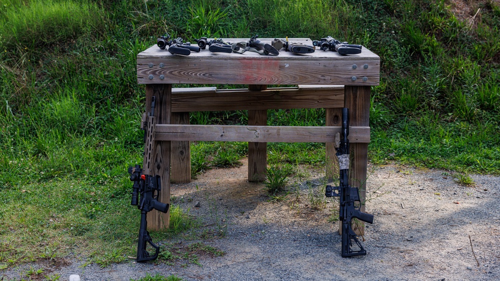 A wooden shooting range table is shown outdoors with two AR-style rifles leaning against its legs on either side. Multiple optics and scopes are laid out on the table surface, suggesting a firearms evaluation or comparison session. The setting appears to be an outdoor shooting range with a gravel surface and grassy berm in the background.