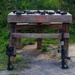 A wooden shooting range table is shown outdoors with two AR-style rifles leaning against its legs on either side. Multiple optics and scopes are laid out on the table surface, suggesting a firearms evaluation or comparison session. The setting appears to be an outdoor shooting range with a gravel surface and grassy berm in the background.